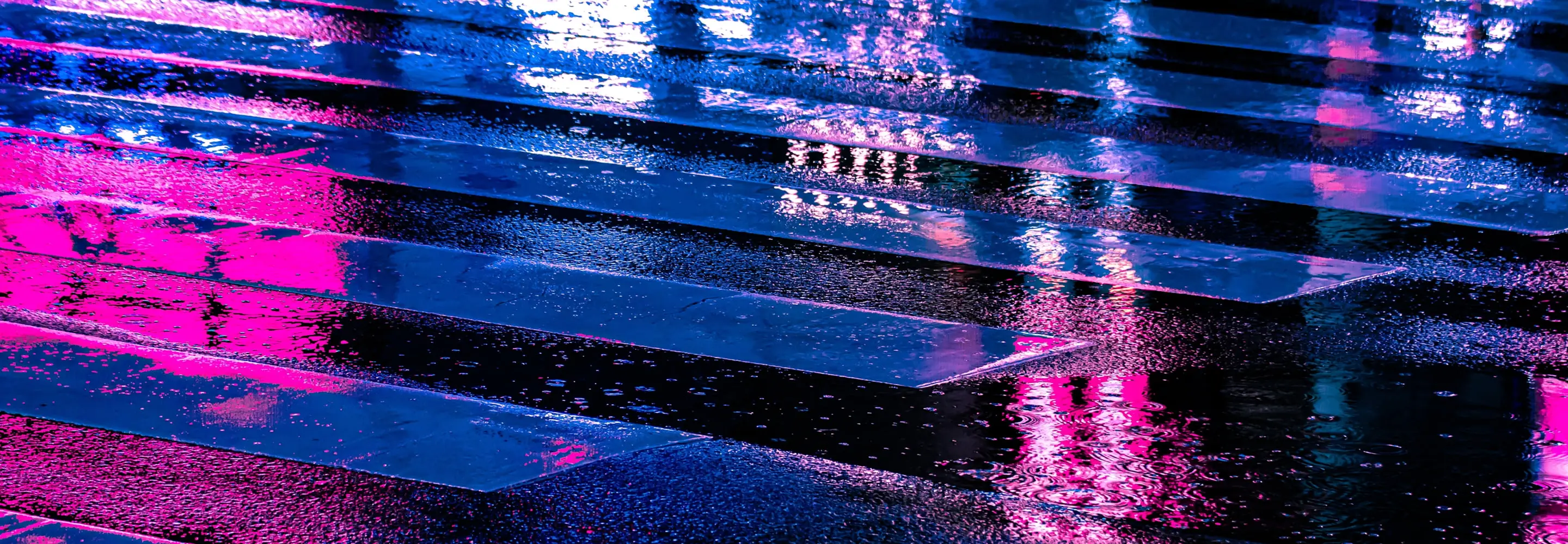青と紫のネオンカラーで照らされた雨の夜の路面の様子。